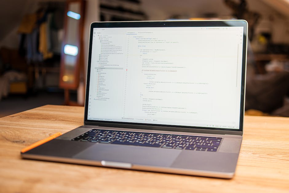A silver laptop displaying lines of code on its screen is placed on a wooden table in a cozy indoor setting with warm lighting. The background is slightly blurred, revealing a bookshelf, a window, and