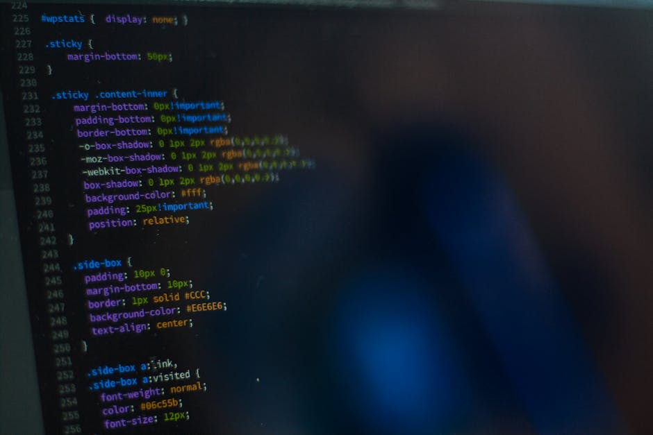A silver laptop displaying lines of code on its screen is placed on a wooden table in a cozy indoor setting with warm lighting. The background is slightly blurred, revealing a bookshelf, a window, and